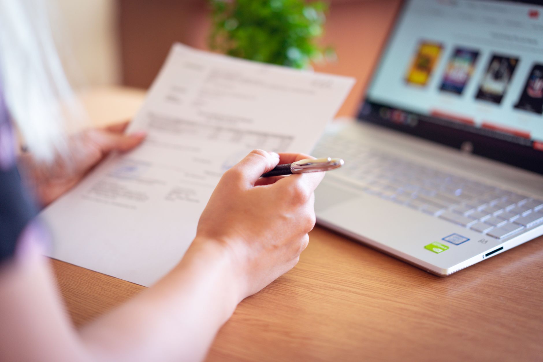 Close-up of a hand holding a pen over a printed document beside an open laptop on a wooden desk