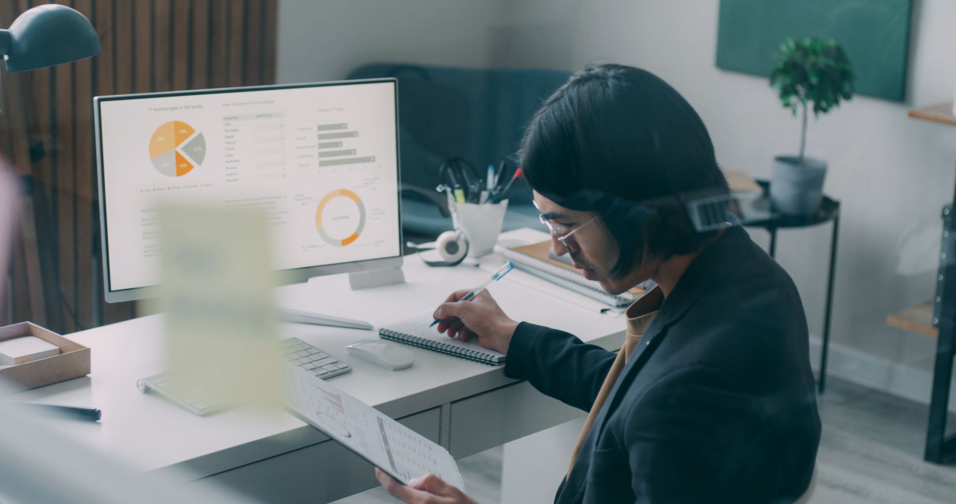 Person at a home desk comparing printed charts with a notebook beside a monitor showing analytics